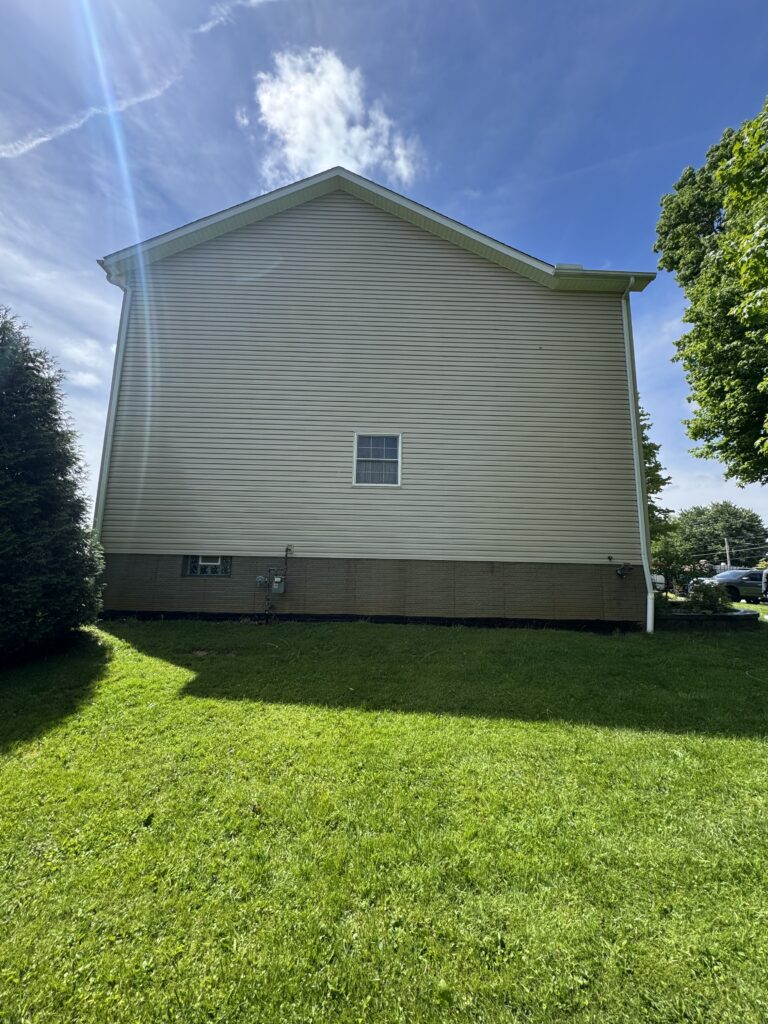 Side view of a beige vinyl-sided house with a small window and a grassy yard under a clear blue sky.