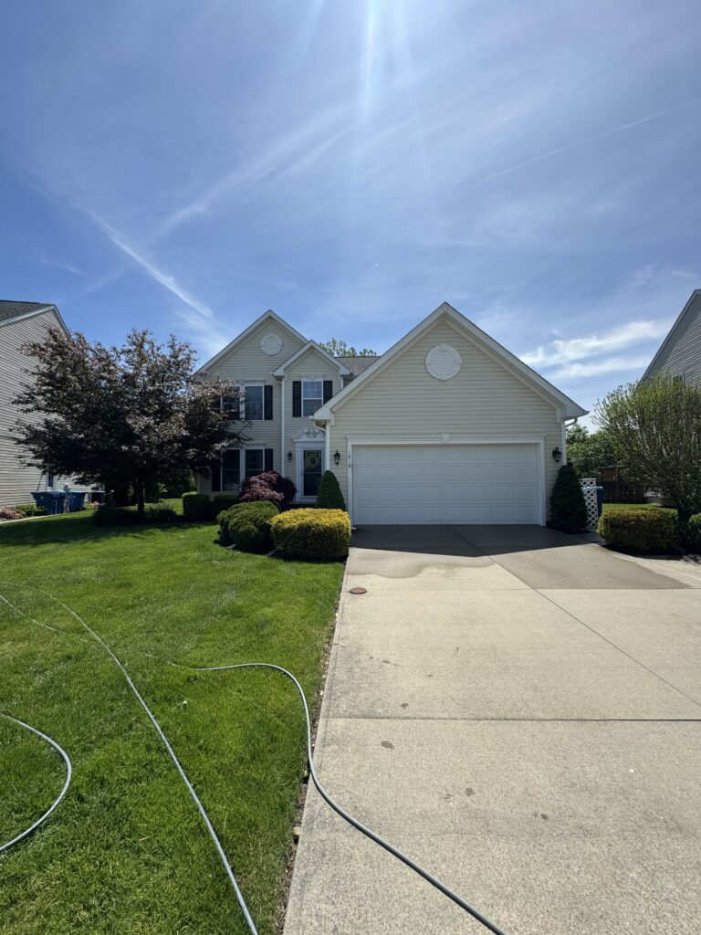 Front view of a beige two-story house with an attached garage, green lawn, and trimmed bushes under a bright blue sky with sun glare, hose on the driveway.