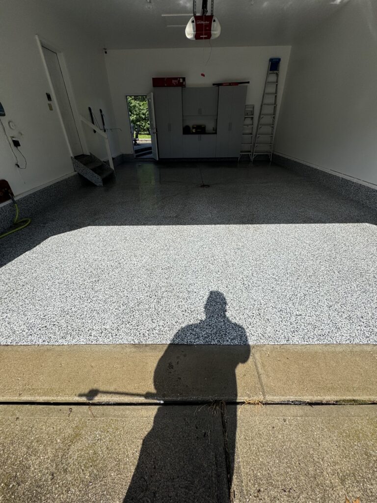 Interior of a clean, empty garage with a speckled epoxy floor, cabinets along the back wall, and a ladder leaning on the right; overhead garage door opener visible.