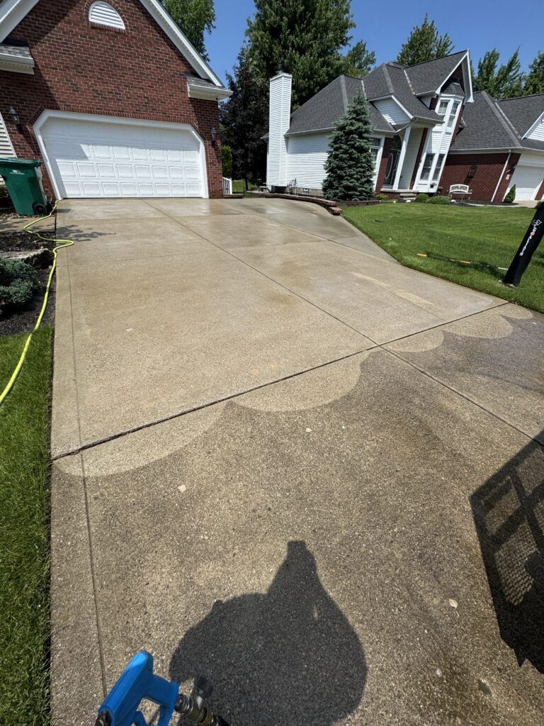 Sunny suburban driveway between brick and white houses, with a large white garage door and a green lawn on the right; yellow hose along the edge and blue equipment in the bottom left corner.
