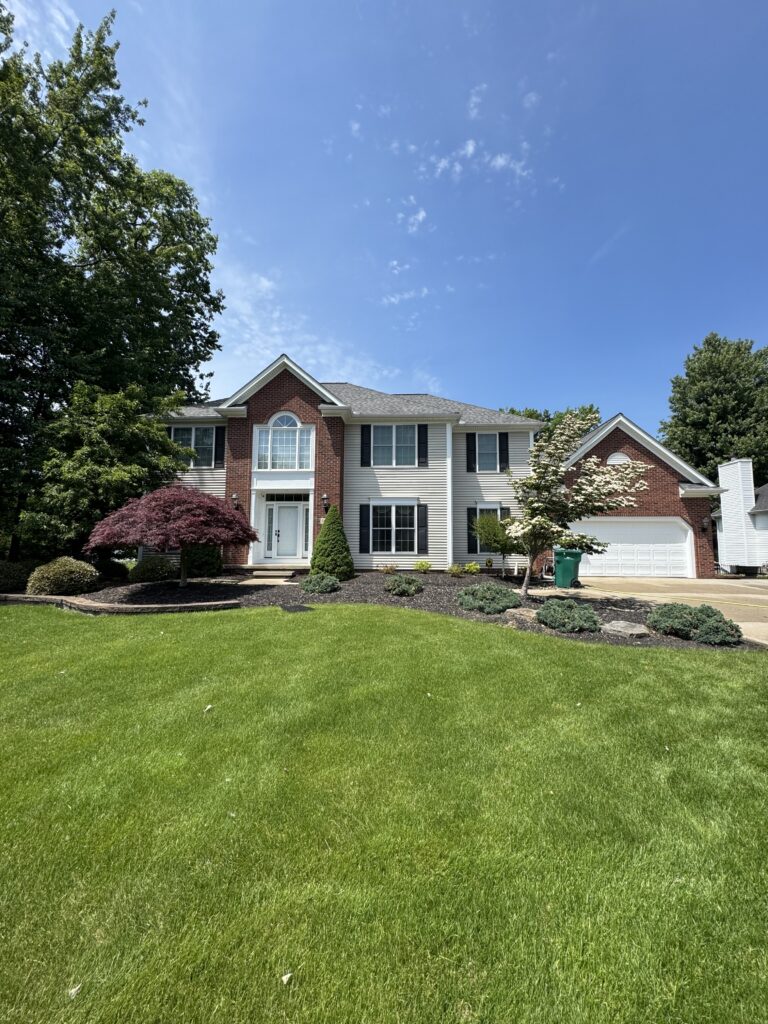 Two-story brick-and-siding suburban house with a manicured lawn and trees under a clear blue sky