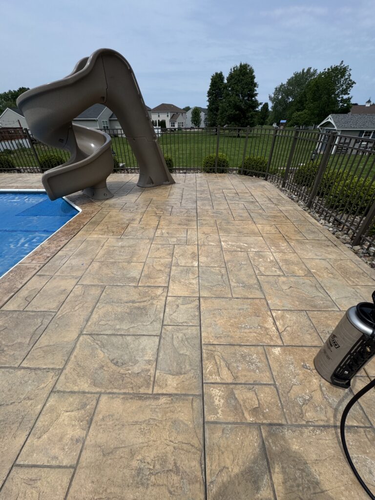 Curved beige backyard pool slide over a tiled patio and blue pool cover, fenced yard beyond.
