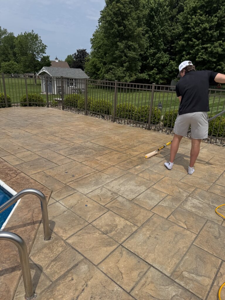 Person in black shirt and white cap standing on a stone patio by a pool, holding a yellow pool tool and looking toward a fenced yard.