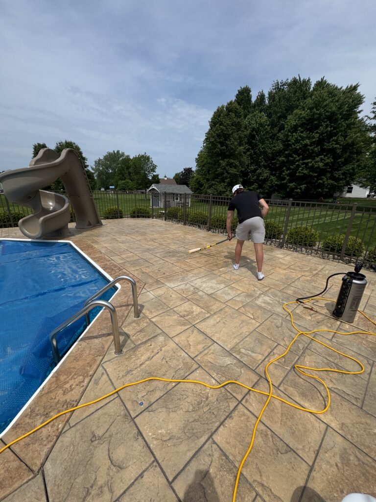 Man in a black shirt and white cap power-washing a tiled pool deck, with a blue pool on the left and a metal fence behind him. A coiled yellow hose runs across the surface. In the background is a grassy yard and large trees.