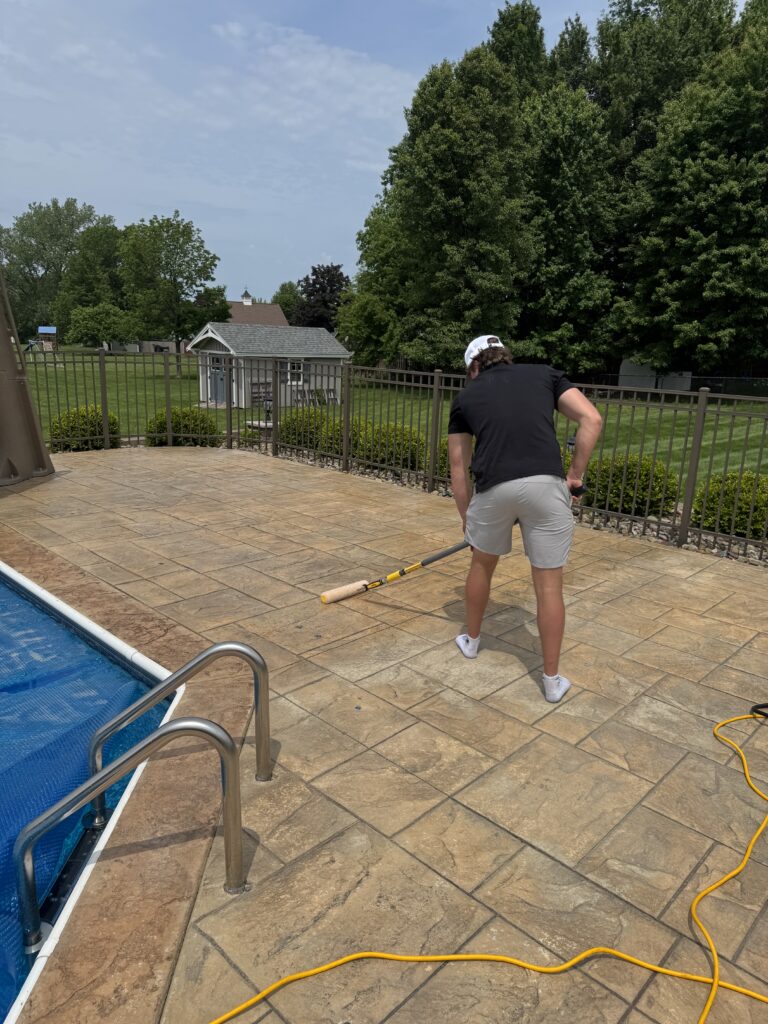 Person in a black shirt and white shorts stands on a tiled pool deck using a long pole to clean the pool edge.