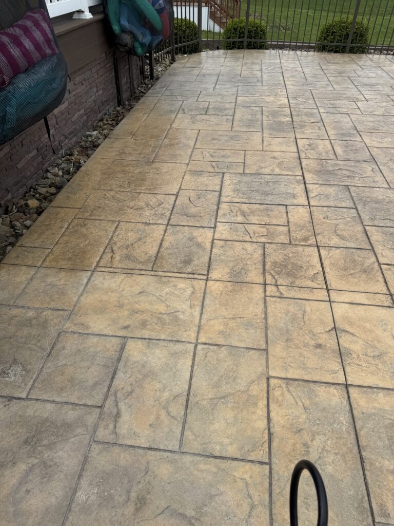Stamped concrete patio with a light tan color and geometric pattern, bordered by a rock bed and brick wall on the left, railing in the distance.
