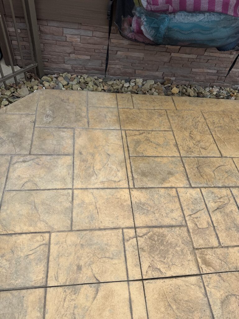 Beige stone patio tiles with a rock edging along a brick wall; a hanging laundry bag is partially visible at the top.