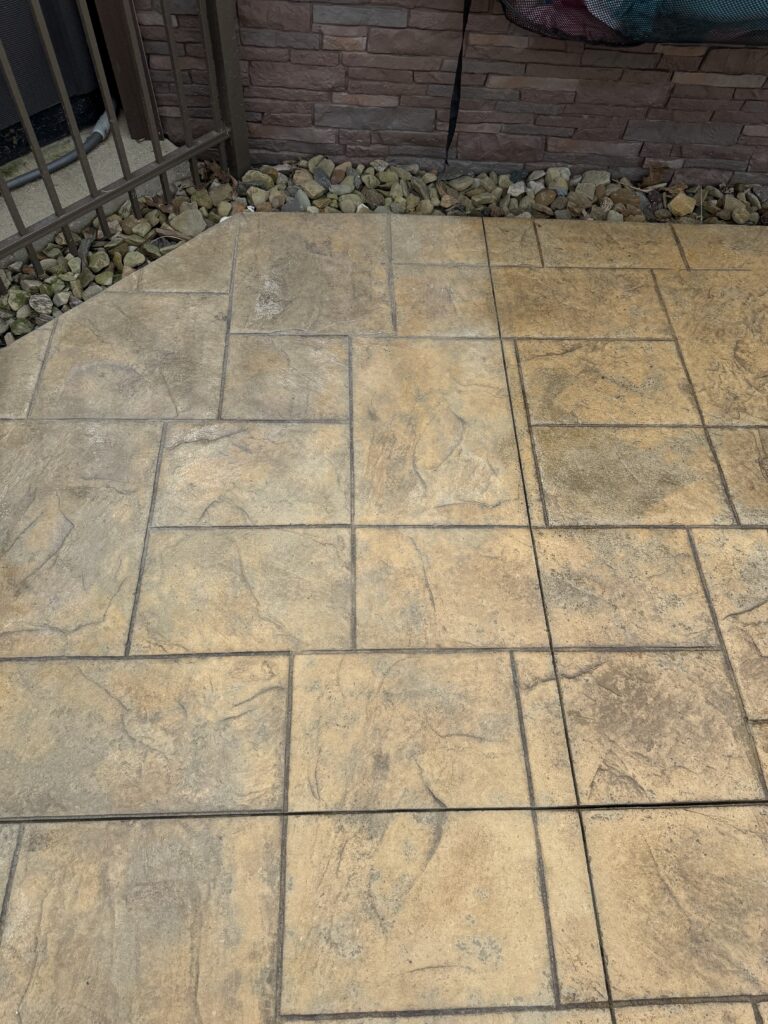 Patio with tan rectangular stone tiles, a rock border along a brick wall, and a metal railing on the left side.