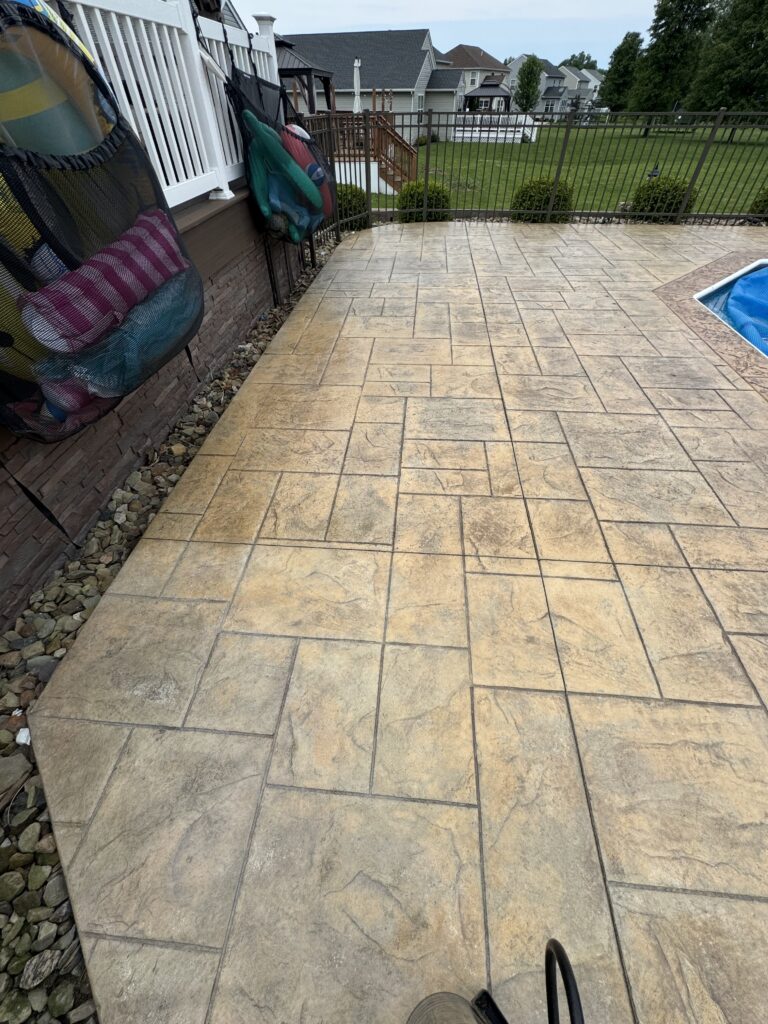 Backyard stamped concrete patio beside the house, with a metal fence and a pool cover visible at the edge.