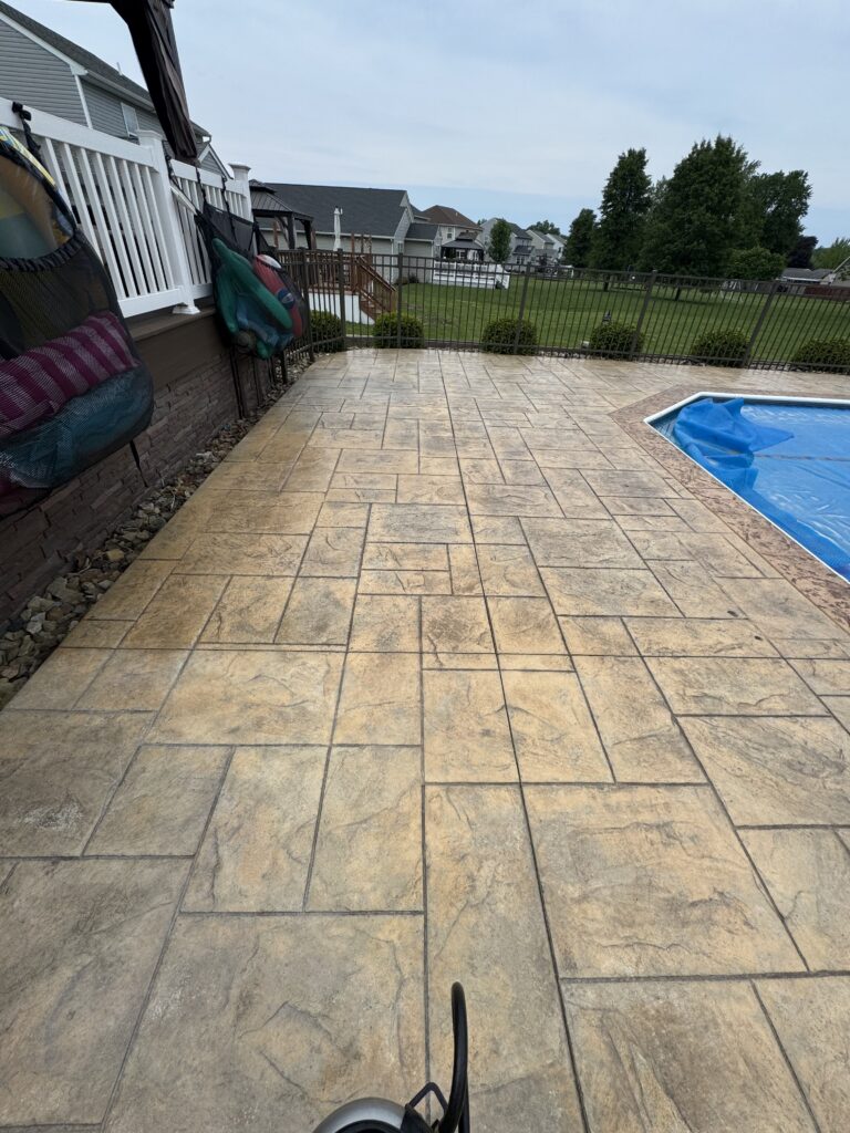 Patio with stamped concrete, black fence, and a covered pool on the right; suburban houses and trees beyond the yard.