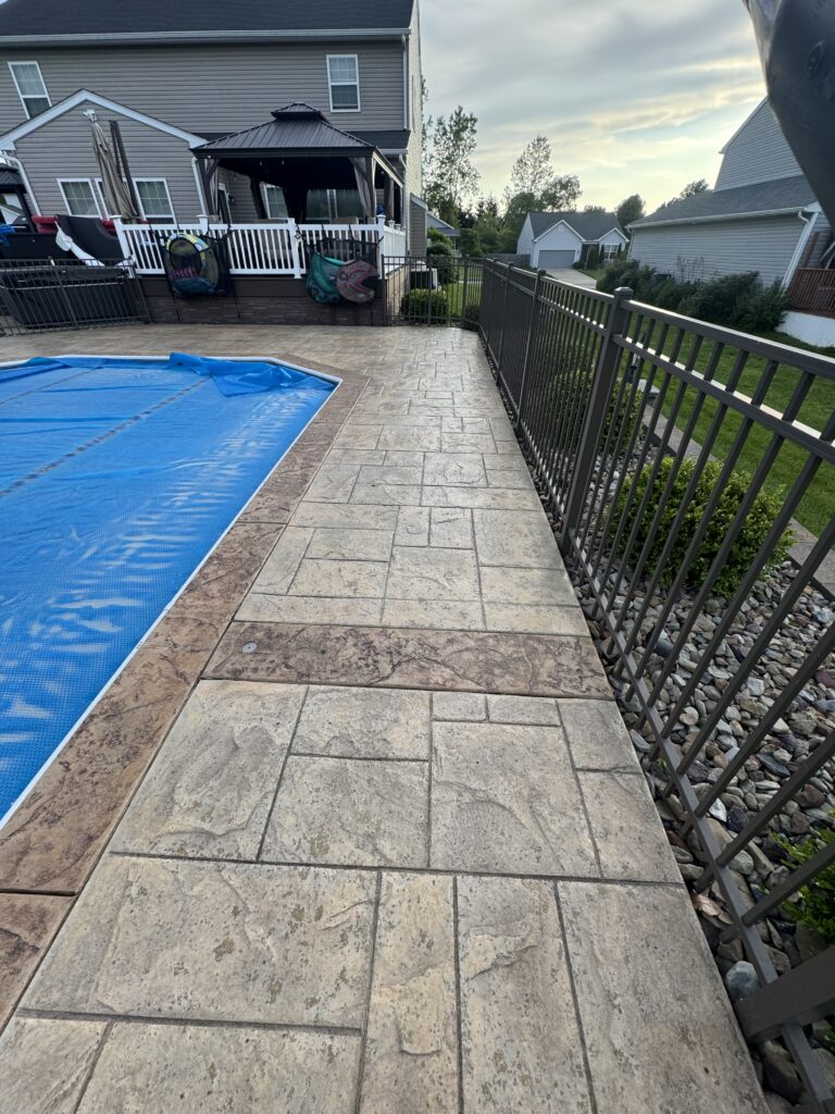 Backyard patio with beige stamped concrete, a blue pool cover on the left, and a black railing along the edge near houses in the background.