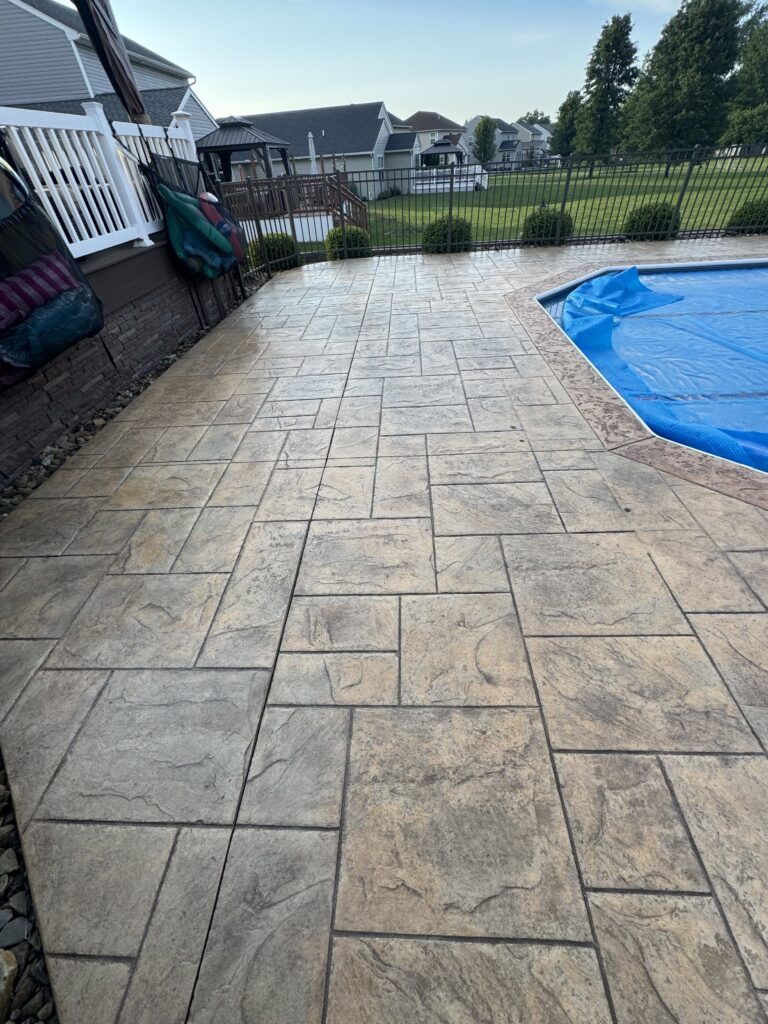 Backyard patio with stamped concrete, black metal fence, and a blue pool cover on the right; suburban houses beyond the lawn.