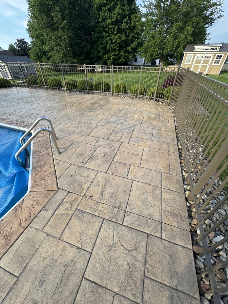 Patio with stamped concrete tiles and a beige metal railing, pool with blue cover on the left, green lawn and trees beyond the fence.