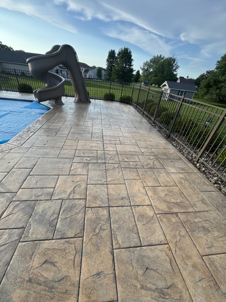 Gray plastic curved pool slide on a beige stone pool deck, with a metal fence and green lawn beyond the yard.