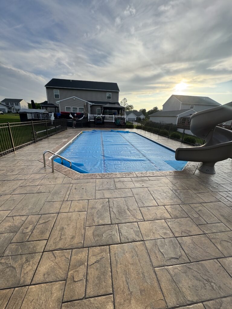 Backyard scene with a rectangular in-ground pool covered by a blue tarp, surrounded by a stone deck and black metal fence.