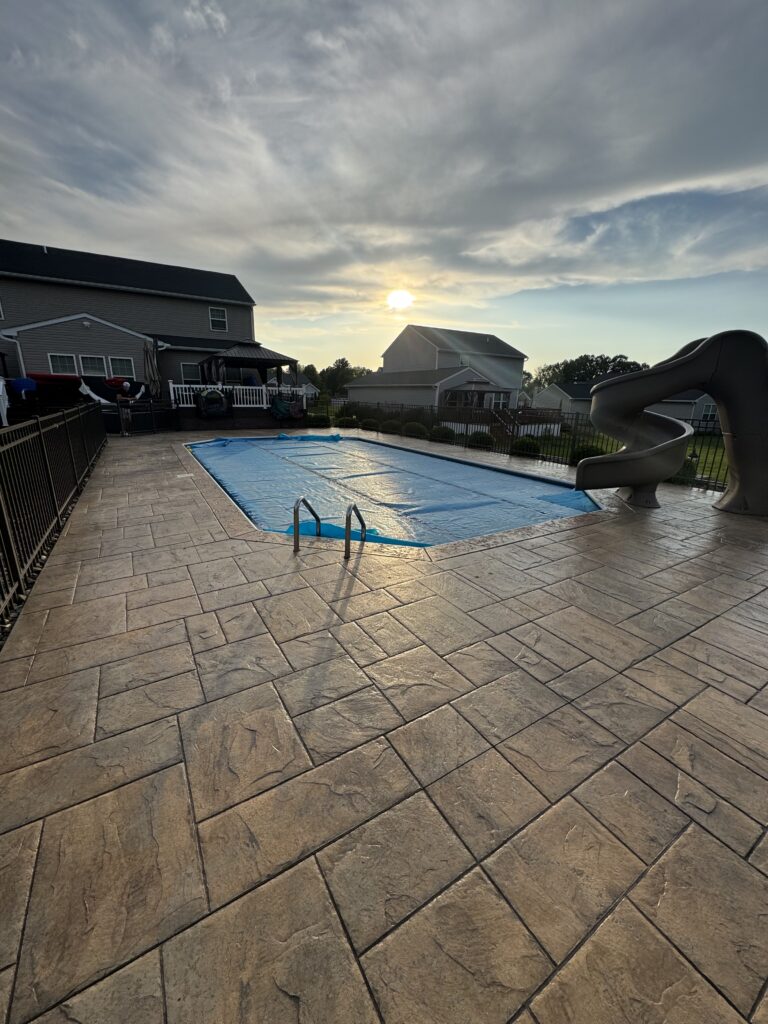 Backyard with an in-ground pool covered by a blue safety cover, tan stone patio, black fence, and neighboring houses; sun near the horizon.