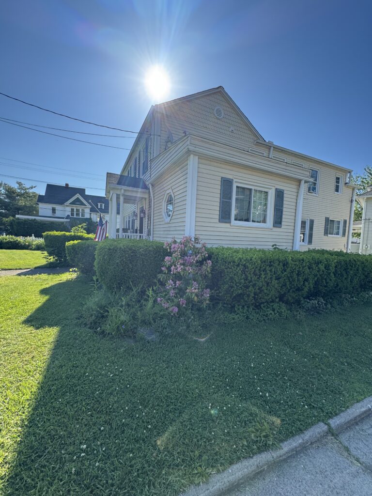 Beige suburban house with blue shutters, a well-kept hedge, and pink flowering bushes under a bright sunny sky.