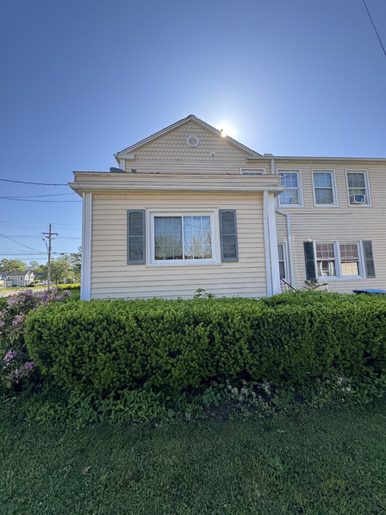 Beige two-story house with a gabled roof, blue shutters, and a full green hedge in front.
