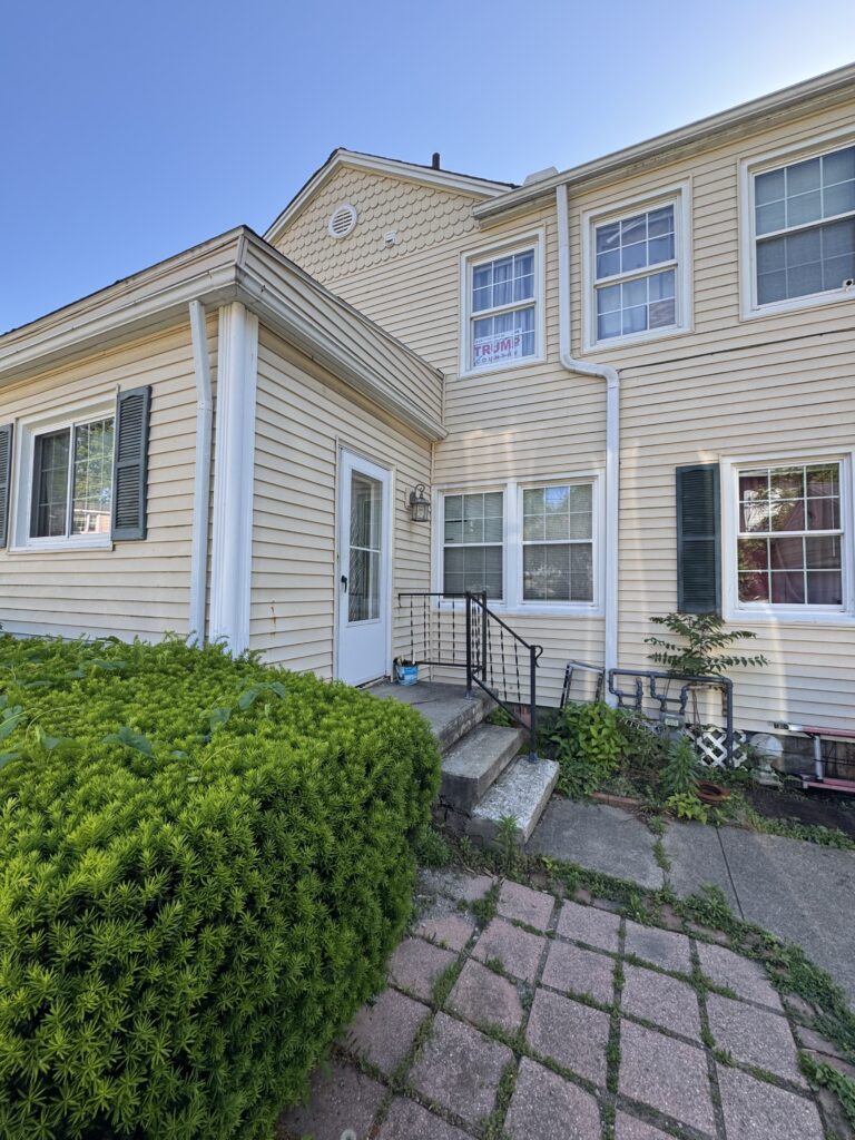 Two-story beige house with white trim, front door and steps, flanked by green shrubs and a stone pathway; a campaign sign visible in a window.
