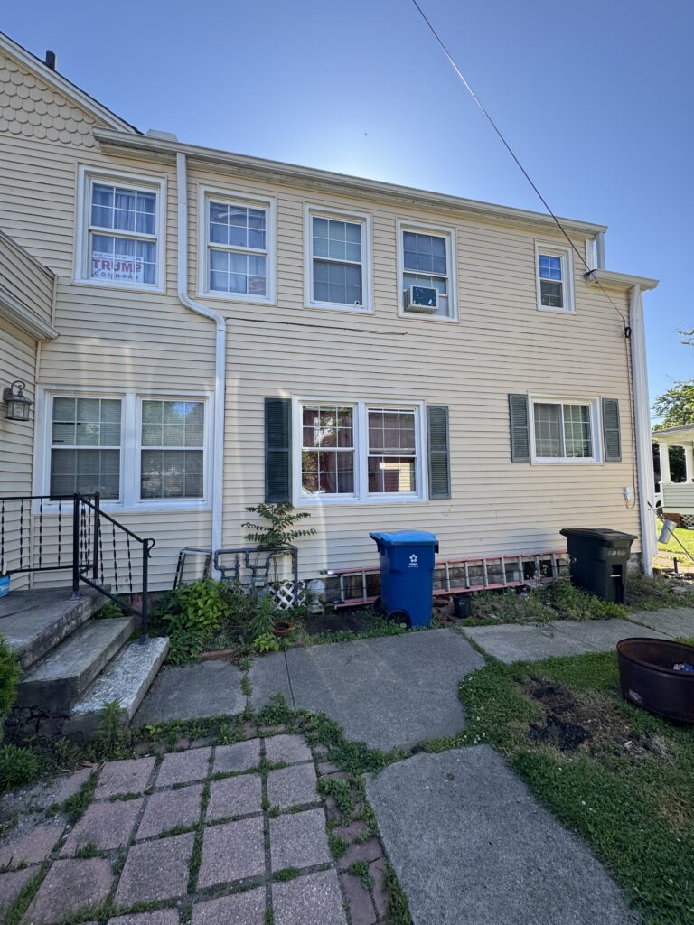 Rear view of a beige siding house with white-framed windows, downspout, and a cluttered yard featuring a blue recycling bin and a black trash bin; a Trump campaign sign is visible in the left window.
