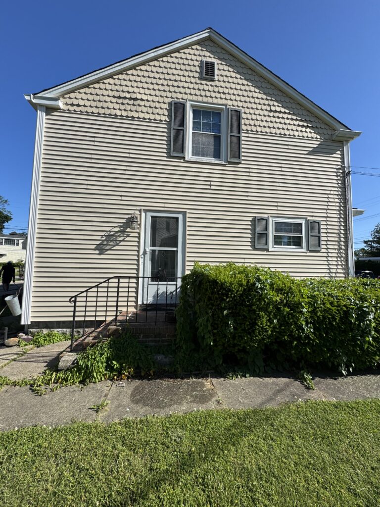 Two-story beige house with scalloped siding on the upper facade, a window with dark shutters, and a small front door under a gabled roof against a blue sky.