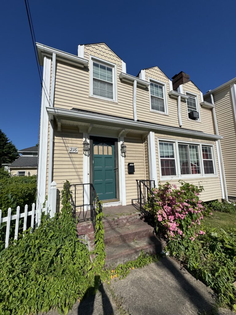 Front view of beige townhouse with teal door, white trim, and floral bushes by a concrete walkway under a clear blue sky