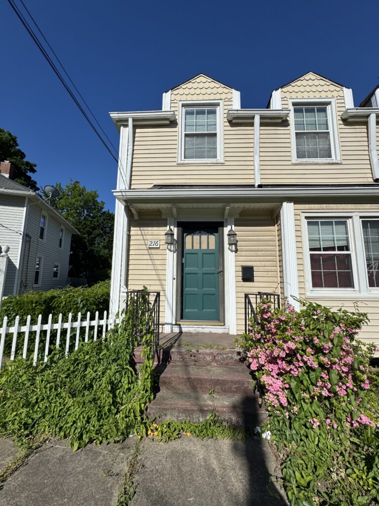 Two-story beige house with green front door, white trim, and two dormer windows under a clear blue sky; pink flowering bush along steps and a white picket fence nearby.