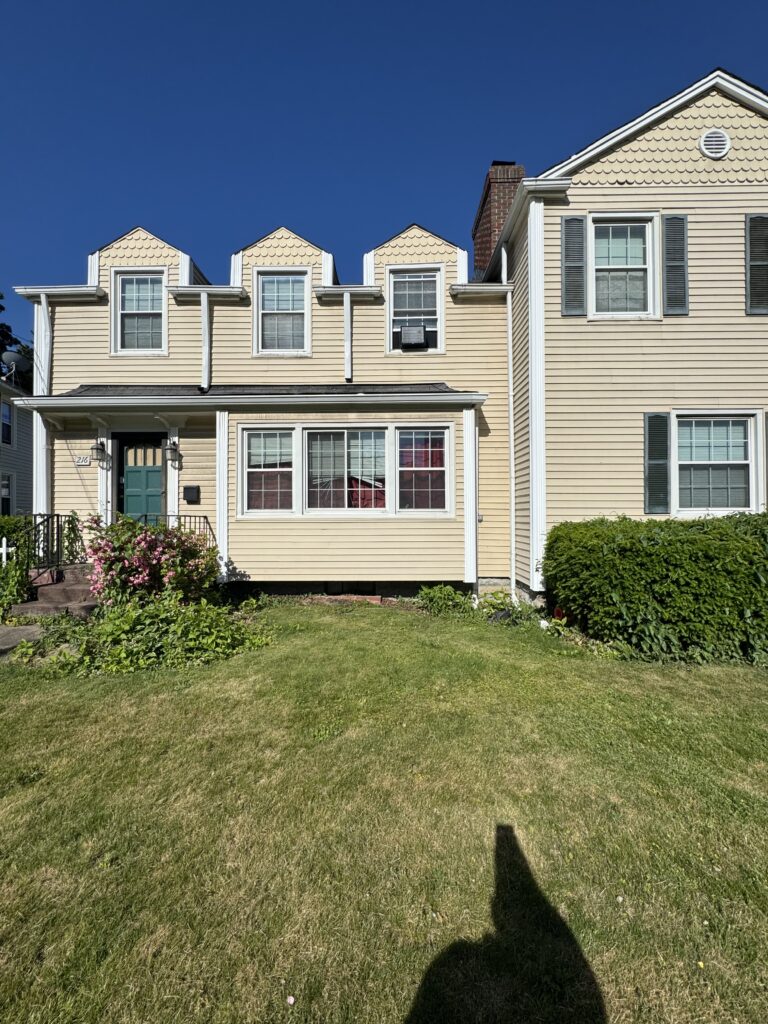 Beige two-story house with dormer windows and a teal front door under a clear blue sky, front yard and shrubs in view.