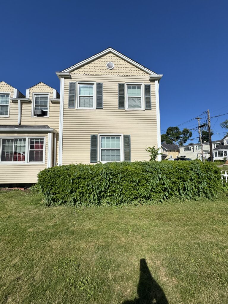 Beige townhouse with green hedge in front and a clear blue sky; two-story home with white trim and shutters.