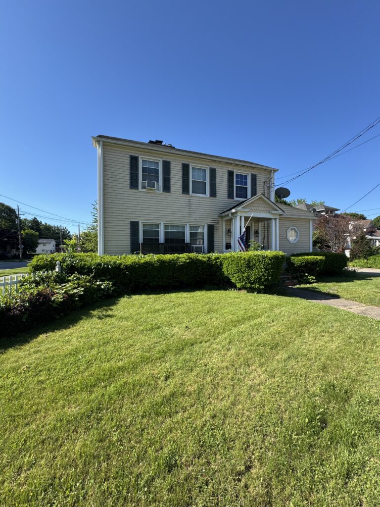 Two-story beige house with dark shutters, small front porch, and an American flag in a well-kept yard under a bright blue sky.