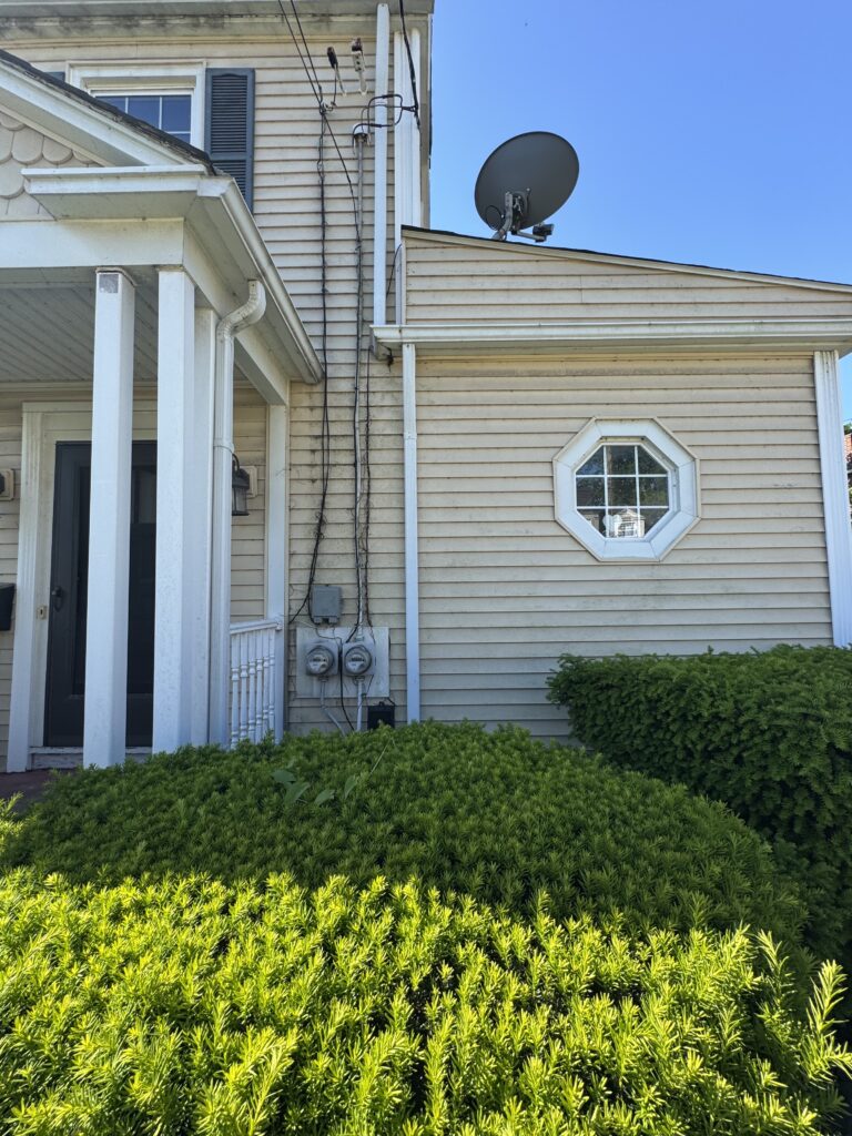Residential house exterior with beige siding, white trim, and a satellite dish on the roof above a small overhang, plus visible utility meters and wires along the wall.