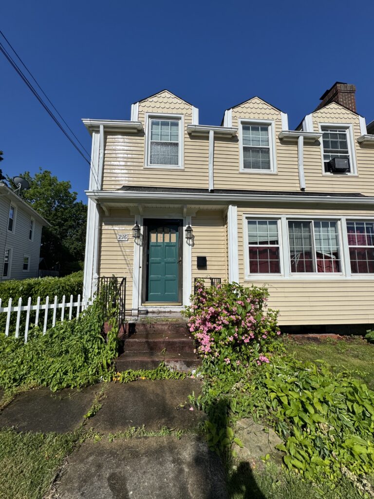 Front view of a beige house with a teal front door, white trim, and pink flowering bushes by the steps under a blue sky.