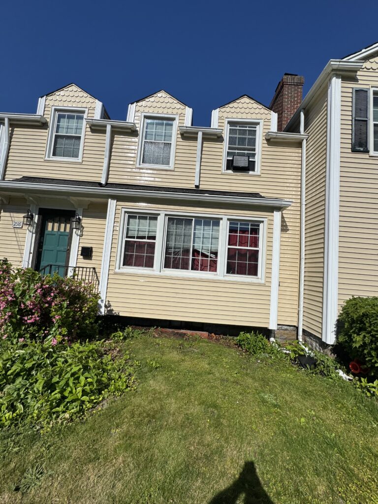 Row of beige townhouses with dormer attic windows, white trim, and a green front door under a clear blue sky, with a flowering shrub in the yard.