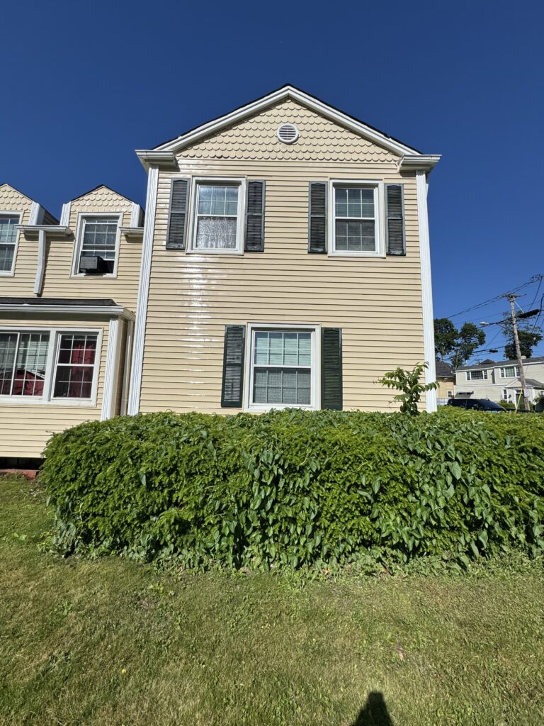 Beige two-story townhouse with black shutters and large windows, a tall green hedge in front, under a clear blue sky