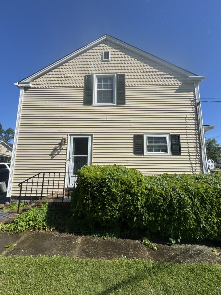 Two-story beige house with decorative fish-scale shingles on the gable, white trim, and black shutters surrounding a central window; front door with steps to the left.