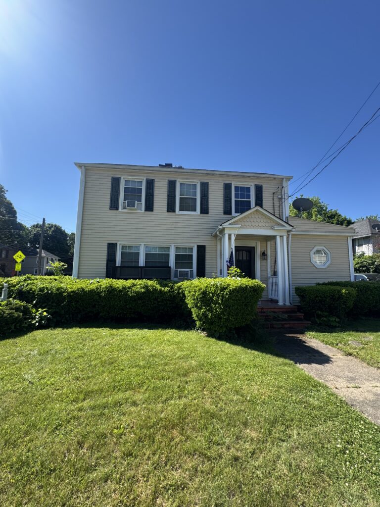 Two-story beige house with black shutters and a small porch, American flag by the entrance, surrounded by a trimmed hedge and green lawn under a clear blue sky