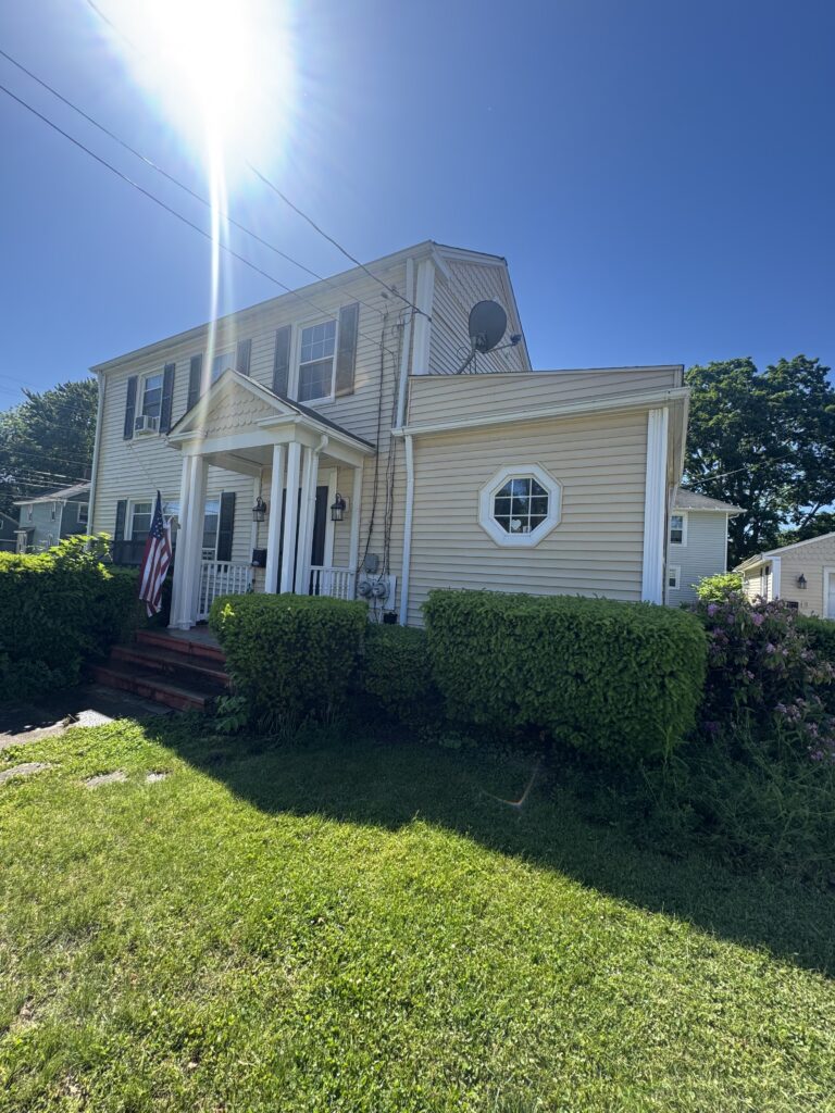 Sunny daytime view of a beige two-story house with a front porch, white trim, and neatly trimmed hedges; American flag near the steps.