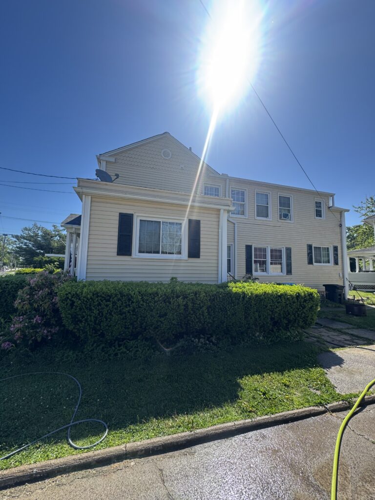 Two-story beige house with white trim and dark shutters under a bright midday sun; a garden hose lies on wet pavement in the foreground.