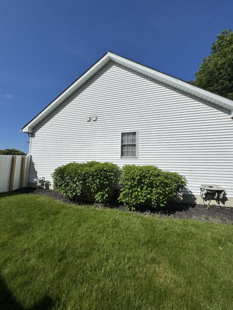 White house side with a steep triangular roof, single window, and trimmed siding, set against a blue sky and shrubs in front.