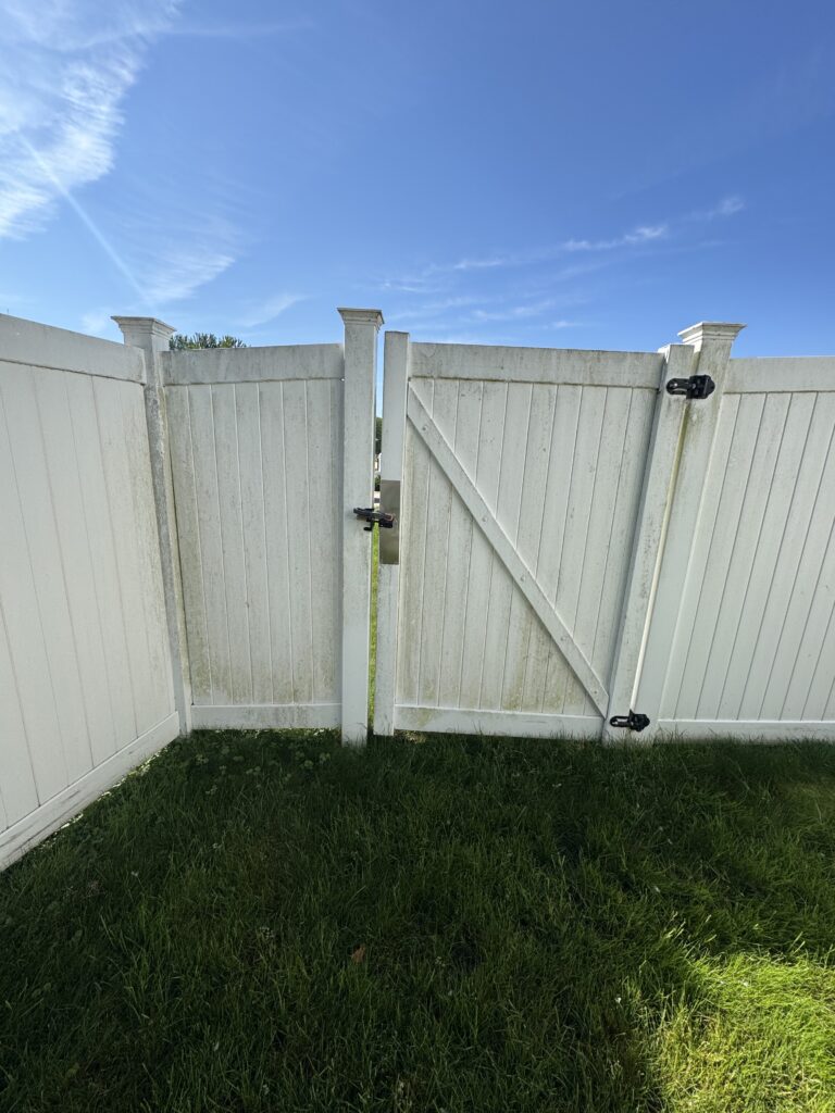 White wooden fence with a slightly open gate set against a clear blue sky and green lawn below.