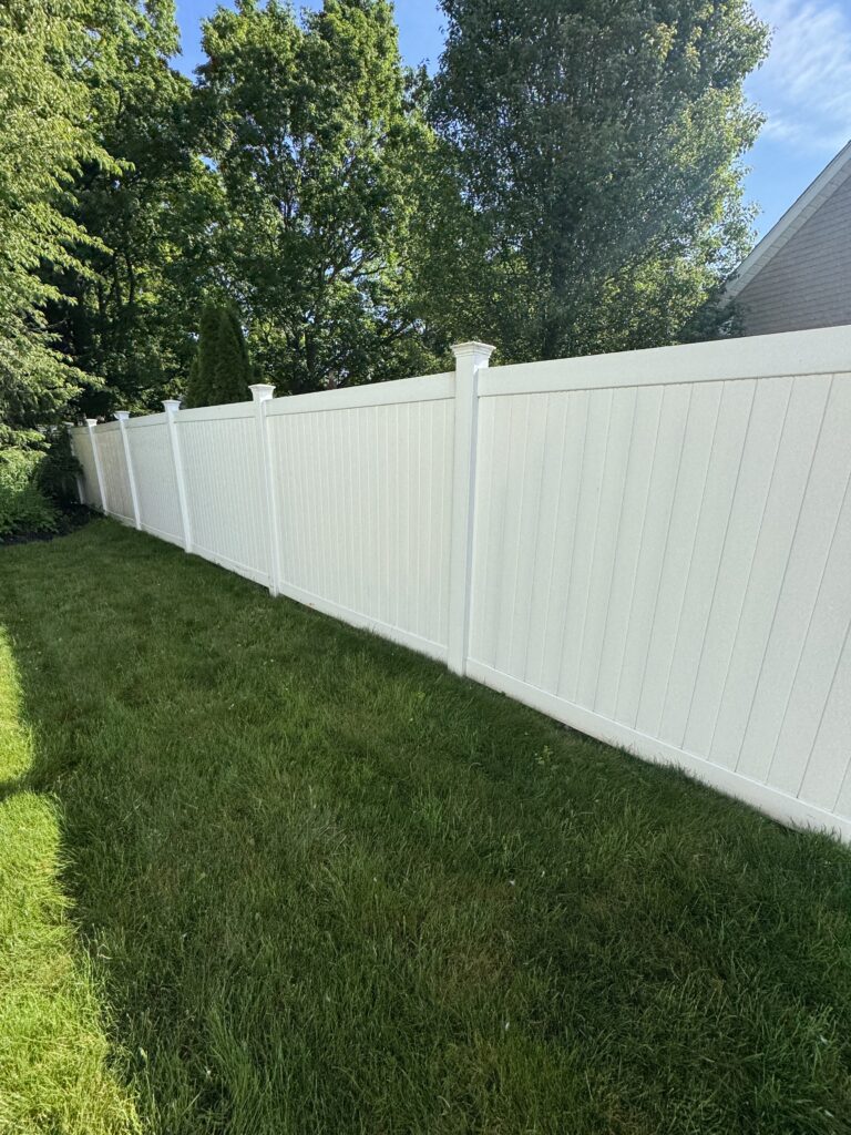 White vinyl privacy fence runs along a lush green lawn with tall trees and blue sky beyond.