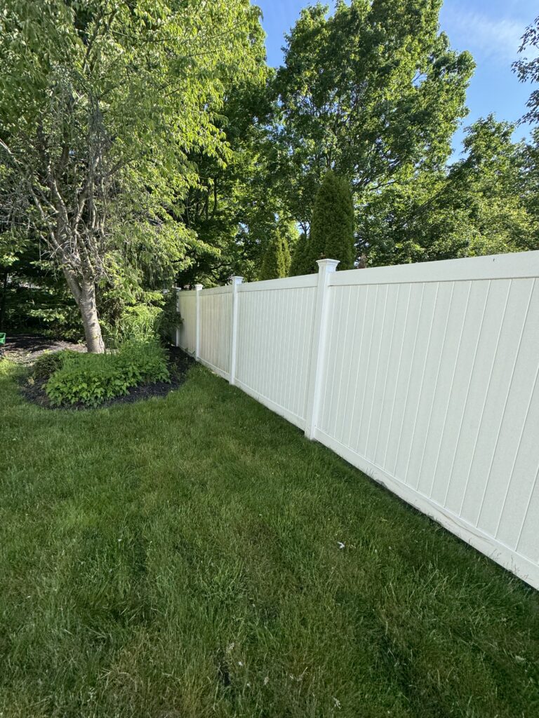 Backyard with a white privacy fence, green lawn, and tall trees along the edge, under a blue sky.