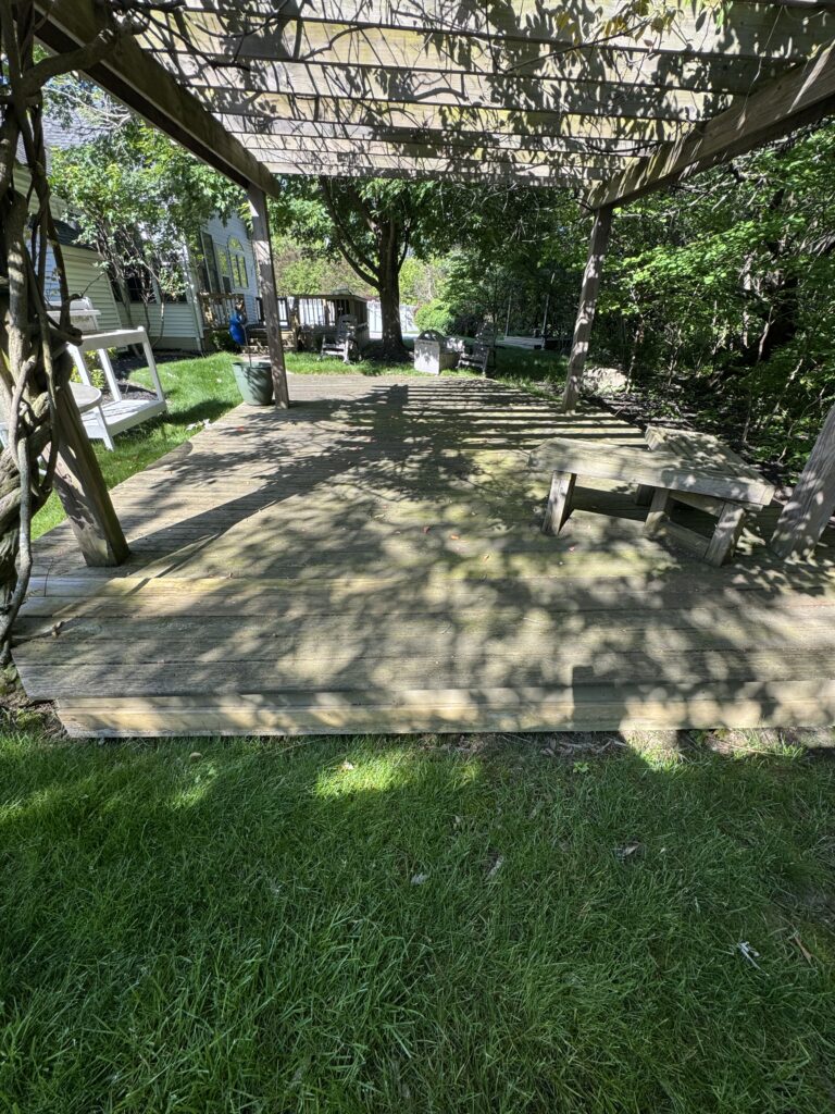 Wooden backyard deck under a trellis, with seating and surrounding green lawn and trees