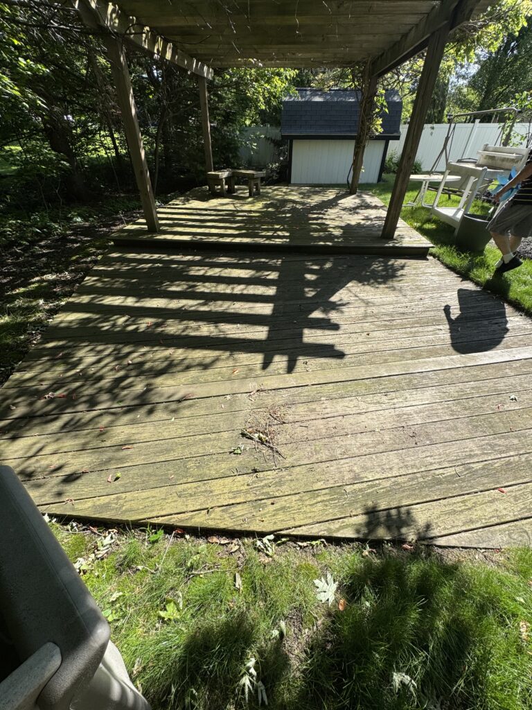 Wooden backyard deck under a pergola with shadows, a small shed in the background, and greenery surrounding the area.