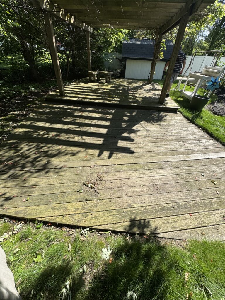 Sunlit wooden backyard deck beneath a pergola, with a man in a striped shirt walking along the right and a shed in the background.