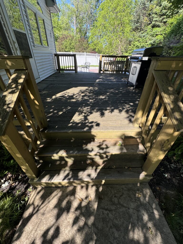 Wooden back deck with stairs, railing, and a gas grill on the right; shade from trees and a fenced backyard beyond.