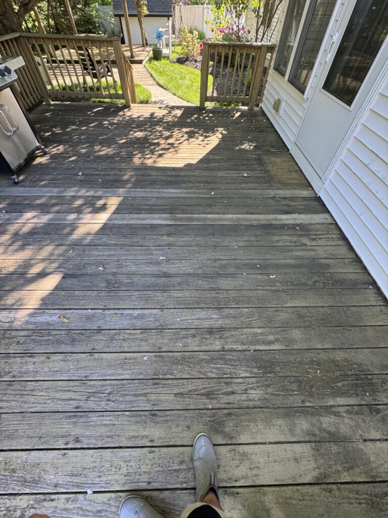 From a first-person perspective standing on a weathered wooden deck in sandals, looking toward a backyard with steps, railing, and a garden. A grill is visible on the left.