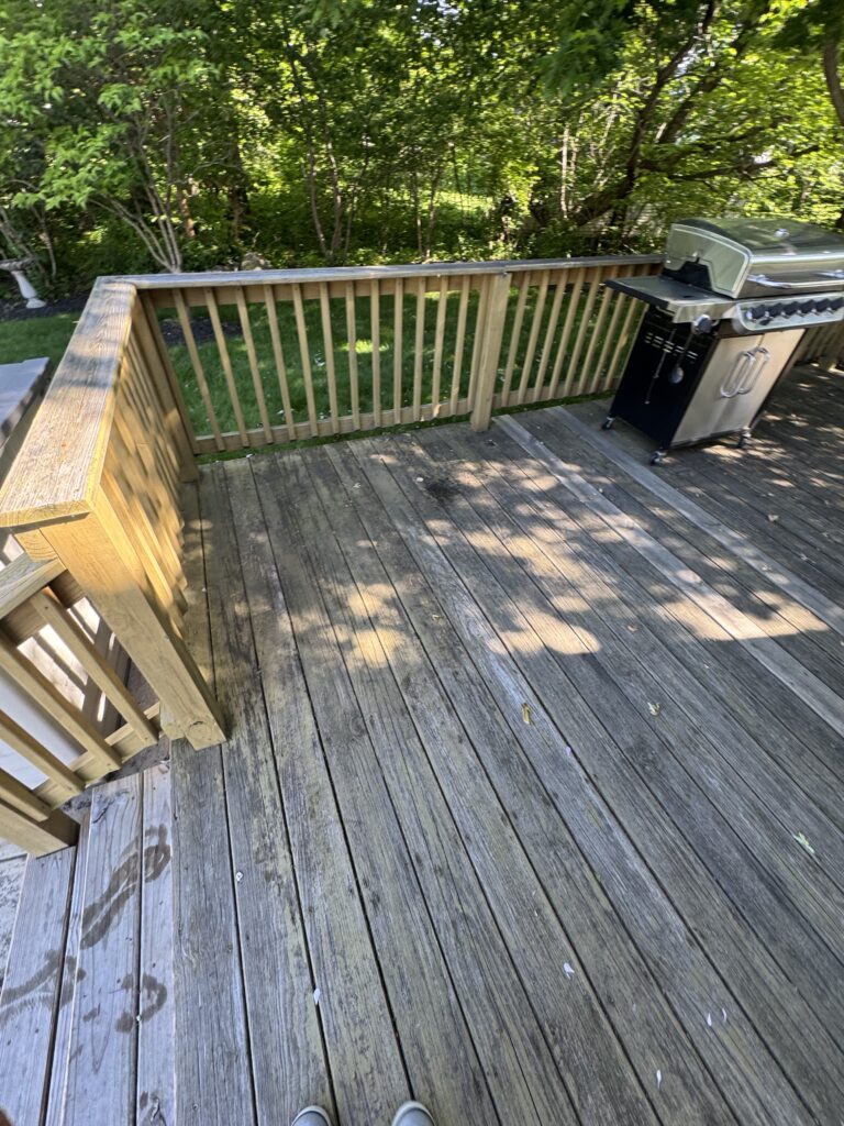 Perspective from a wooden deck showing a stainless gas grill on the right, railing along the edge, and dense green trees beyond; shoes at the bottom edge indicate viewer position.