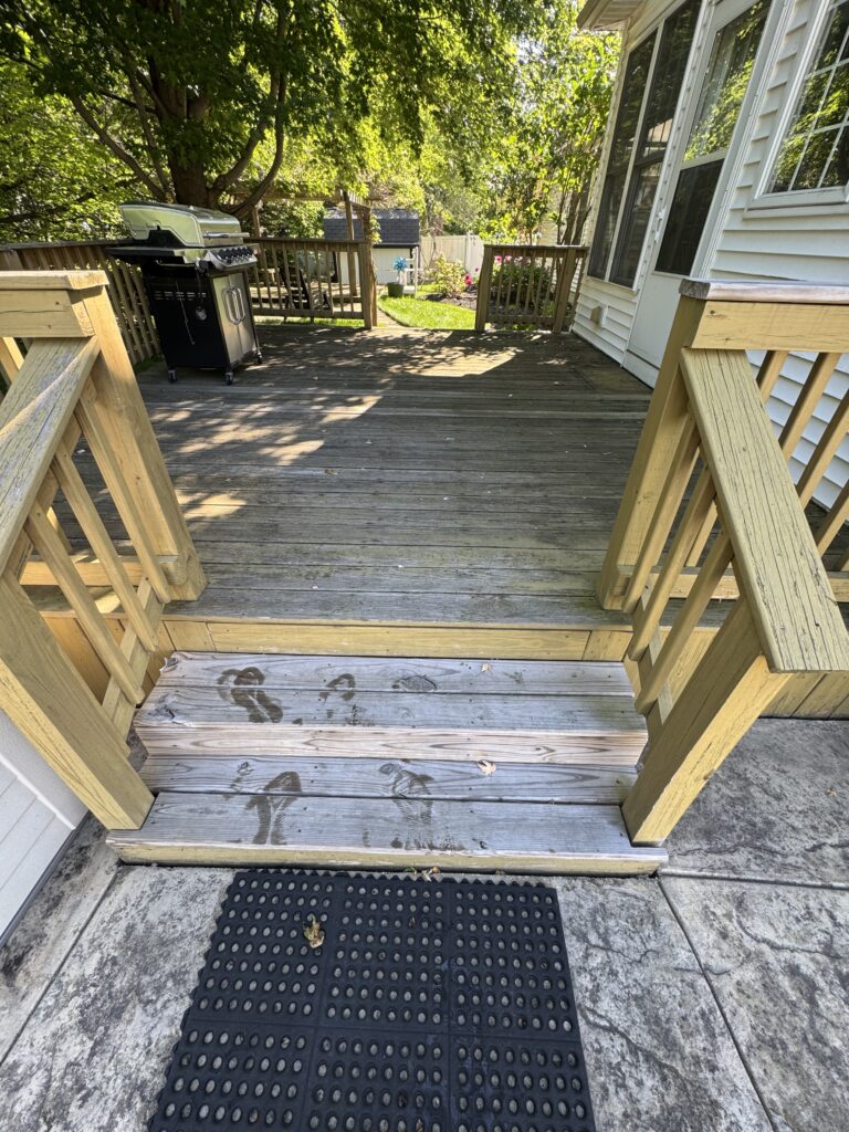 Gas grill on a wooden deck next to a sliding door, with a sunny backyard and trees beyond the railing.