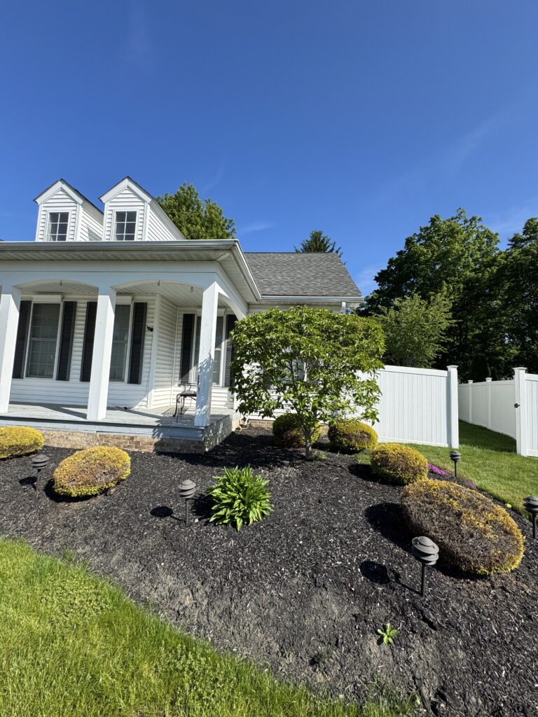 White siding house with a covered front porch, dormer windows, and a manicured black-mulch garden with trimmed shrubs under a clear blue sky.